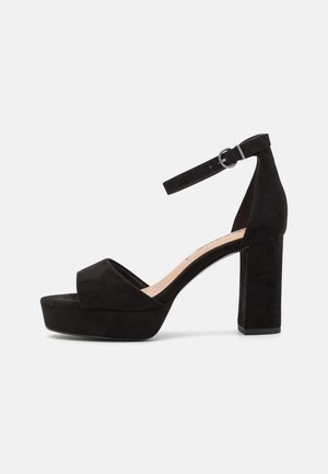 Sort ruskind platform sandal med blokhæl, ankelrem og åben tå, vist fra siden på en ensfarvet baggrund.