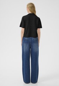 Femme aux cheveux blonds portant un haut noir à manches courtes et un jean bleu ample taille haute, se tenant de dos sur un fond blanc.