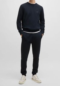 Tmavě modrá mikina a odpovídající tepláky z jemné látky, s malým logem na hrudi a bílým detailem na pasu, doplněné teniskami.