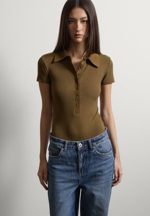 Junge Frau trägt ein eng anliegendes, olivgrünes, geripptes Poloshirt mit kurzen Ärmeln und eine hoch taillierte blaue Jeans, steht vor einem schlichten Hintergrund.