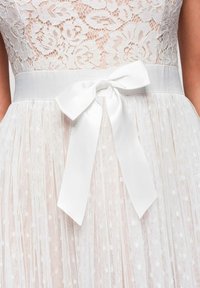 Robe blanche en dentelle avec motif floral sur le corsage, jupe en tulle transparent à pois et large ceinture en satin nouée en nœud à la taille.