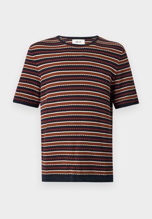 Homme portant un haut en maille rayé à manches courtes, un pantalon beige à jambes larges et des sandales marron foncé, debout avec une main dans la poche sur un fond uni.