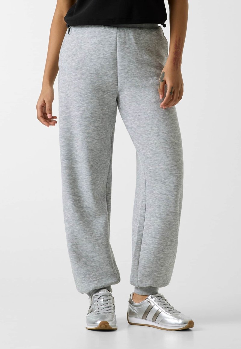 Personne portant un pantalon de survêtement gris clair et des baskets argentées, debout devant un fond uni, mains détendues le long du corps.