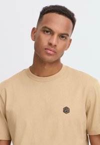 Lyhythihainen beige T-paita, joka on valmistettu pehmeästä kankaasta. Vasemmassa rinnassa on pieni musta geometrinen brodeerattu logo. Pyöreä kaula-aukko.