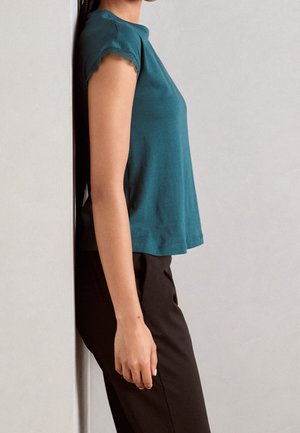 Personne appuyée contre un mur gris clair portant un haut turquoise à manches courtes et un pantalon noir, debout de côté avec le bras relaxé vers le bas.