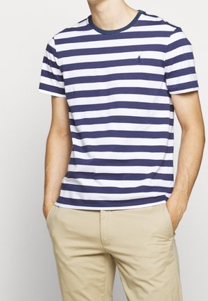 Mann trägt marineblaue und weiße horizontal gestreifte T-Shirt mit Rundhalsausschnitt und beige Hose, Hände in den Taschen.