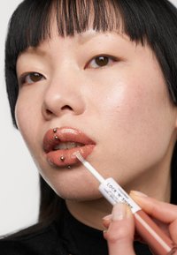 Lipglossrør med en hvid applikator holdt nær glossy, nude-farvede læber udsmykket med piercinger. Glat, blank finish på huden.