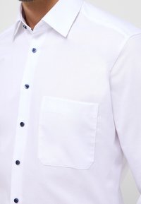 Camisa de vestir blanca con una textura sutil, con cuello puntiagudo, un bolsillo en el pecho y detalles en botones azules.