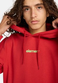 Červená mikina s kapucňou, obsahujúca klokanie vrecko, kapucňu so šnúrkou a malým zeleným logom s nápisom "element" na prednej strane. Mäkká textúra látky.