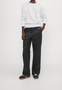 Sudadera blanca con cuello redondo y logo dorado "AX", combinada con pantalones anchos de cuero sintético negro de talle alto y zapatillas negras con cordones blancos.