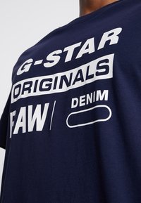 Marinblå bomullströja med iögonfallande vit text: "G-STAR ORIGINALS FAW / DENIM." Korta ärmar och rundad halsringning, mjuk textur.