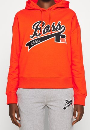Osoba ubrana w jaskrawo pomarańczową bluzę z kapturem z napisem "Boss Athletic" oraz szare spodnie dresowe z dopasowanym logo "Boss Athletic" na udzie.
