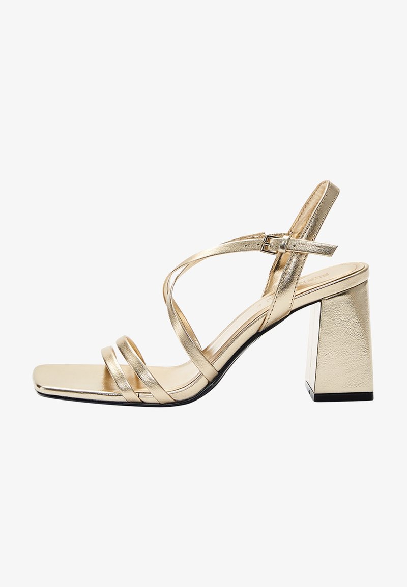 Sandale à talon haut en métal doré avec un bout carré, dotée de lanières croisées et d'un talon carré épais. Texture lisse et détails minimalistes.