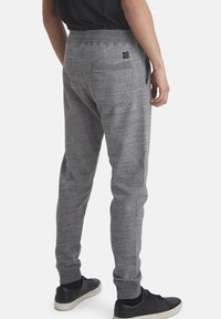 Pantalones deportivos grises con un acabado texturizado, cinturilla elástica y bolsillos laterales, combinados con zapatillas negras. Diseño minimalista con etiqueta de logo.