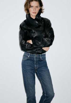 Femme aux cheveux courts portant une veste en cuir noire et un jean bleu, debout devant un fond clair uni, les bras croisés.