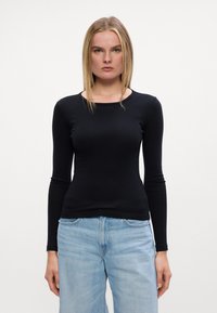 Fekete, hosszú ujjú felső sima anyagból, kör alakú nyakkivágással, világoskék farmernadrággal párosítva. Egyszerű dizájn, testhezálló fazon.