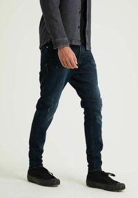 Jean en denim bleu foncé avec une coupe slim, présentant un léger effet usé et un design classique à cinq poches, associés à des baskets noires.