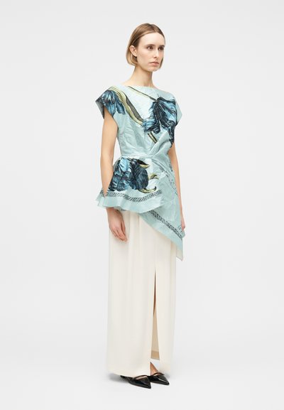 Γυναίκα που φοράει ασύμμετρη ανοιχτό μπλε μπλούζα με λουλουδένιο μοτίβο, με draped σχεδιασμό και αμάνικο, συνδυασμένη με μακρύ λευκό φούστα και μαύρα μυτερά παπούτσια.