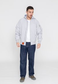 Graue Bomberjacke mit Kapuze, Fleece-Futter und gerippten Bündchen, getragen über einem weißen Hemd und dunklen Jeans, mit einem Logopatch.