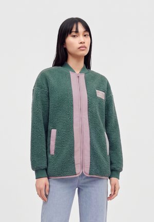 Jeune femme aux longs cheveux noirs portant une veste en polaire verte avec des bordures roses et un jean bleu clair sur un fond uni clair.