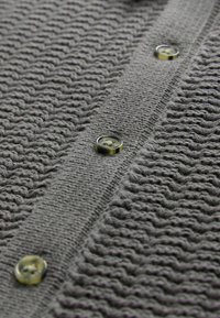Cardigan en tricot gris avec un motif zigzag texturé, fermeture à boutons avec des boutons translucides verts et une finition douce et duveteuse.