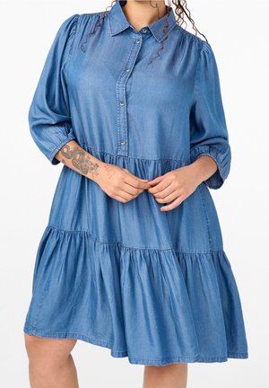 Kobieta ubrana w jasnoniebieską, zapinaną na guziki, długorękawową sukienkę dżinsową z wielowarstwową spódnicą, z tatuażem na lewym przedramieniu, z rękami splecionymi na talii.