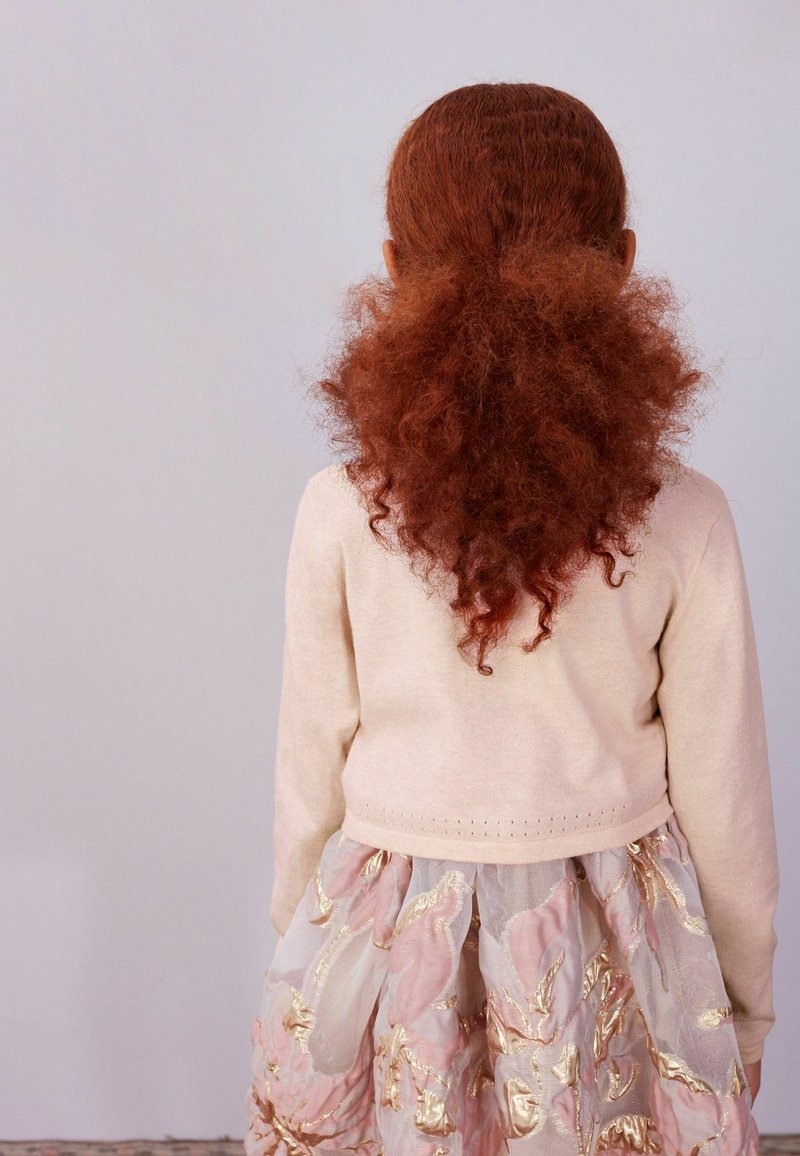 Jeune personne avec de longs cheveux roux bouclés attachés à l'arrière, portant un pull beige et une jupe fleurie rose et or, dos au spectateur.
