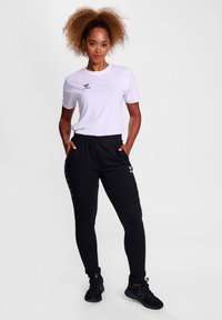Hvid T-shirt med korte ærmer og sort logo, parret med sorte joggingbukser med sidelister og snor i taljen. Sorte sneakers.