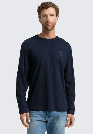Marineblaue Langarmshirts mit rundem Hals und subtiler Logodruck auf der Brust, kombiniert mit hellblauen Jeans. Weicher, strukturierter Stoff.