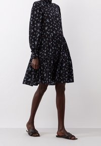Robe noire à fleurs avec de longues manches bouffantes, décolleté haut et ourlet froncé. Présente de petites fleurs bleues ; fabriquée en tissu léger. Sandales noires.