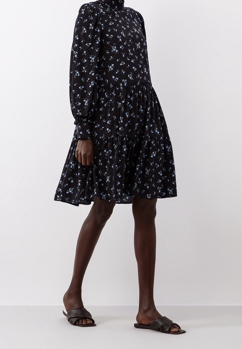 Robe noire à fleurs avec de longues manches bouffantes, décolleté haut et ourlet froncé. Présente de petites fleurs bleues ; fabriquée en tissu léger. Sandales noires.