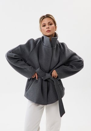Femme portant un manteau ceinturé gris foncé oversize avec un col montant et un pantalon blanc, debout devant un fond clair uni.