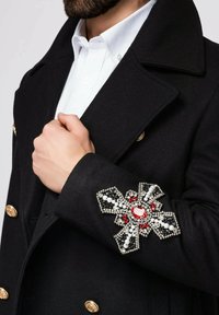 Schwarzer Wollmantel mit goldenen Knöpfen und einer dekorativen Brosche am Ärmel, die aus Perlen sowie roten, weißen und schwarzen Steinen besteht.