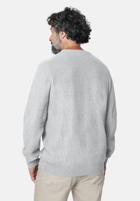 Hombre con cabello rizado y barba, vistiendo un suéter de punto gris claro y pantalones beige, mostrado desde la espalda frente a un fondo blanco liso.