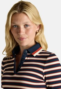 Blondīne ar gaišām freckles, ģērbusies zeltainās riņķa auskaros un tumši zili, sarkani un baltā svītrainā polo krekliņā, mierīgi skatās kamerā.