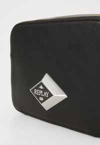 Svart läderpärm med en silverfärgad, diamantformad metallplatta som har texten "REPLAY 1981" på framsidan.