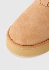 Beige suede kingad ümara varbaga, millel on tekstureeritud pealis ja pruun kummist tall. Serva ääristav õmblus lisab detaili.