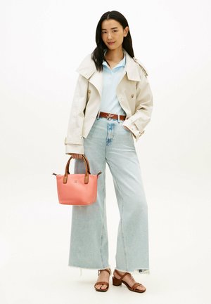 Vrouw draagt een beige trenchcoat, lichtblauwe polo, wijde jeans, bruine sandalen, en houdt een kleine roze handtas met bruine handvaten vast.