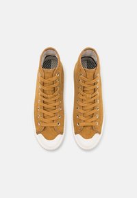Brązowe korduroyowe sneakersy za kostkę z białymi gumowymi podeszwami, wyposażone w metalowe oczka i płaskie sznurowadła, widok z góry podkreślający teksturowaną tkaninę.
