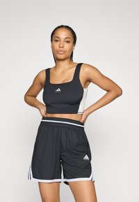Kvinne i svart og hvit atletisk crop-top og løse basketballshorts som står med hendene på hoftene mot en ensfarget bakgrunn.