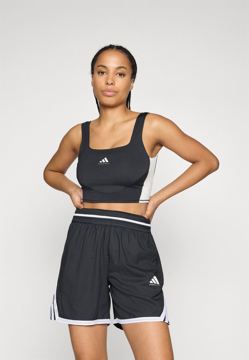 Kvinne i svart og hvit atletisk crop-top og løse basketballshorts som står med hendene på hoftene mot en ensfarget bakgrunn.