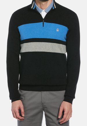 Hombre con jersey negro con rayas horizontales azules y grises y cuello con cremallera hasta media altura, combinado con pantalones grises.