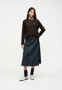 Γυναίκα που φοράει μακρυμάνικο μαύρο τοπ, μπεζ ζιβάγκο, midi φούστα από σκούρο τζιν, μαύρες ζαρωμένες κάλτσες και λευκά loafers σε λευκό φόντο.