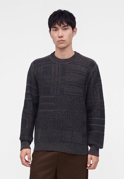 Jeune homme aux cheveux noirs courts portant un pull en maille texturé foncé et un pantalon marron, debout devant un fond clair uni.