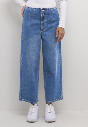 Bredbenta blå denimbyxor med hög midja, plisseringar, knäppning framtill och fransade fållar, kombinerade med vita sneakers.