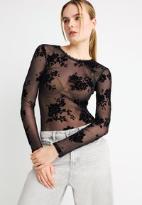 Μαύρο μακρυμάνικο τοπ από δίχτυ με βελούδινη κεντητική λουλουδιών, στρογγυλή λαιμόκοψη και κυματιστές άκρες, συνδυασμένο με ανοιχτό γκρι τζιν.