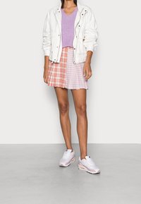 Bílá bunda na zip přes levandulový svetřík s výstřihem do V, doplněná plisovanou sukní v červeném a fialovém kostkovaném vzoru. Na nohou bílé a růžové tenisky.