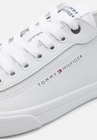 Zapatilla de cuero blanca con cordones blancos, que presenta un logo de TOMMY HILFIGER bordado en el costado y una suela de goma texturizada.