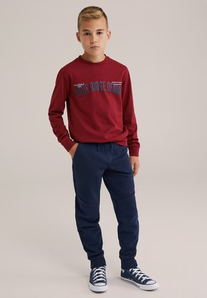 Rode lange mouwen shirt met grafische tekst, gecombineerd met marineblauwe joggingsbroek met kniestukken en marineblauwe sneakers met witte zolen.