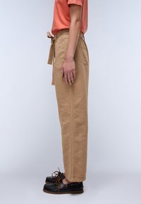 Khaki tapered broeken met een riem in de taille, voorzien van een gladde textuur, zijzakken en een wijde pijpontwerp. Gekleed met zwarte veterschoenen.
